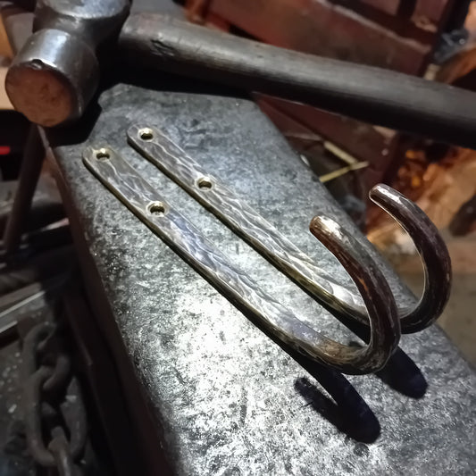 Pair of large hand-forged solid brass wall hooks on a blacksmith's anvil