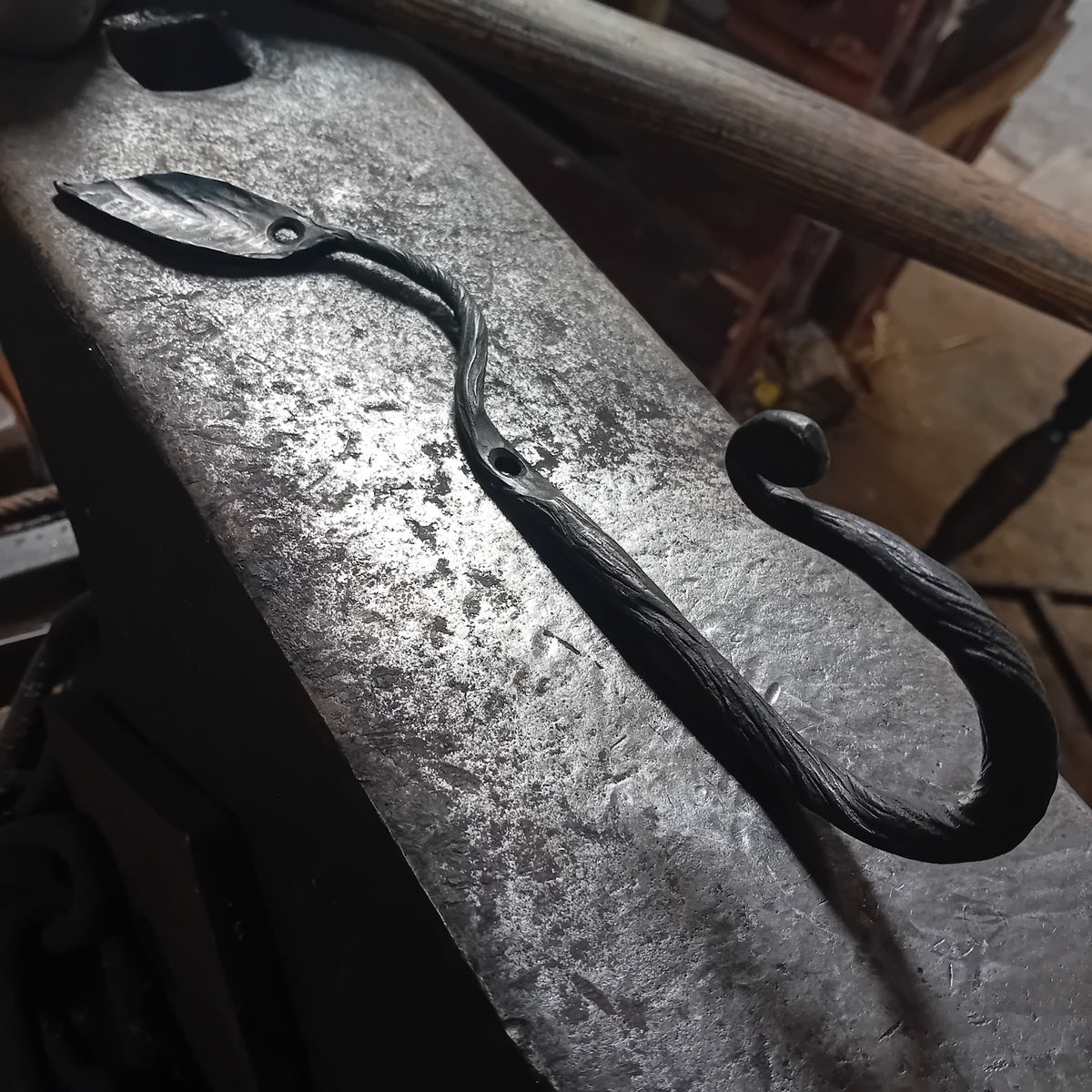 Hand-forged botanical leaf wall hook with twisted stem and scroll end on blacksmith anvil