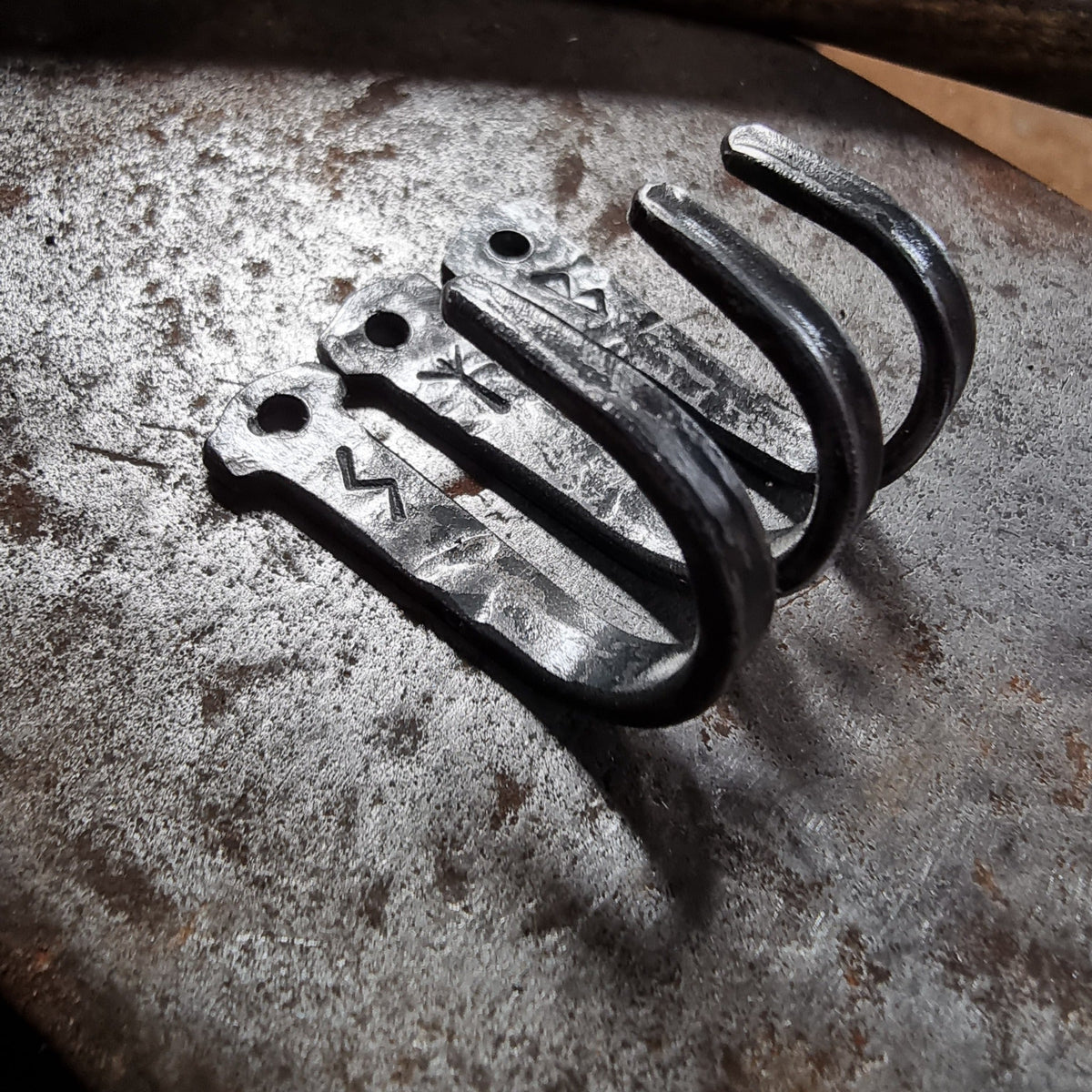 Three hand-forged steel rune hooks with engraved Elder Futhark symbols, photographed on a forge anvil
