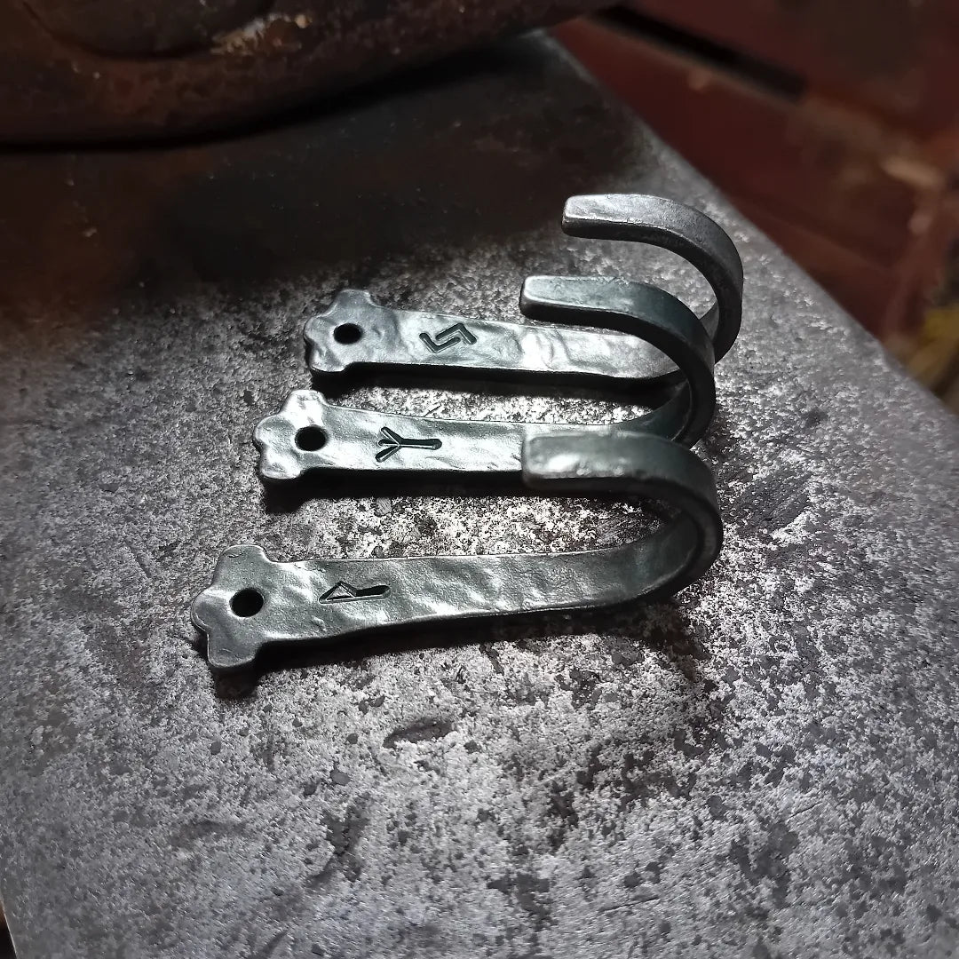 Three hand-forged Elder Futhark rune hooks on a textured anvil, showing engraved runes, fixing holes and curved hooks